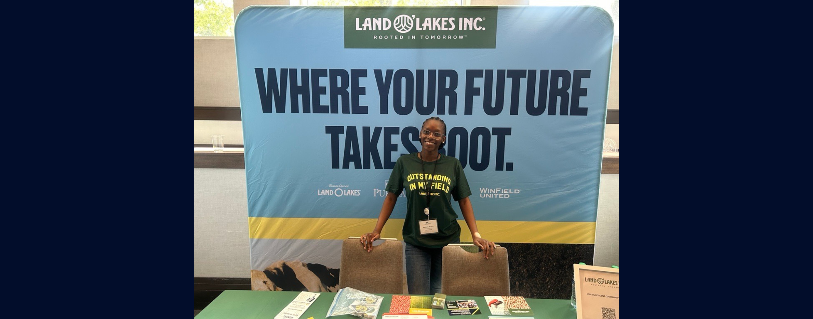 Mekaela stands at a job fair booth with a "Where Your Future Takes Root" banner behind her