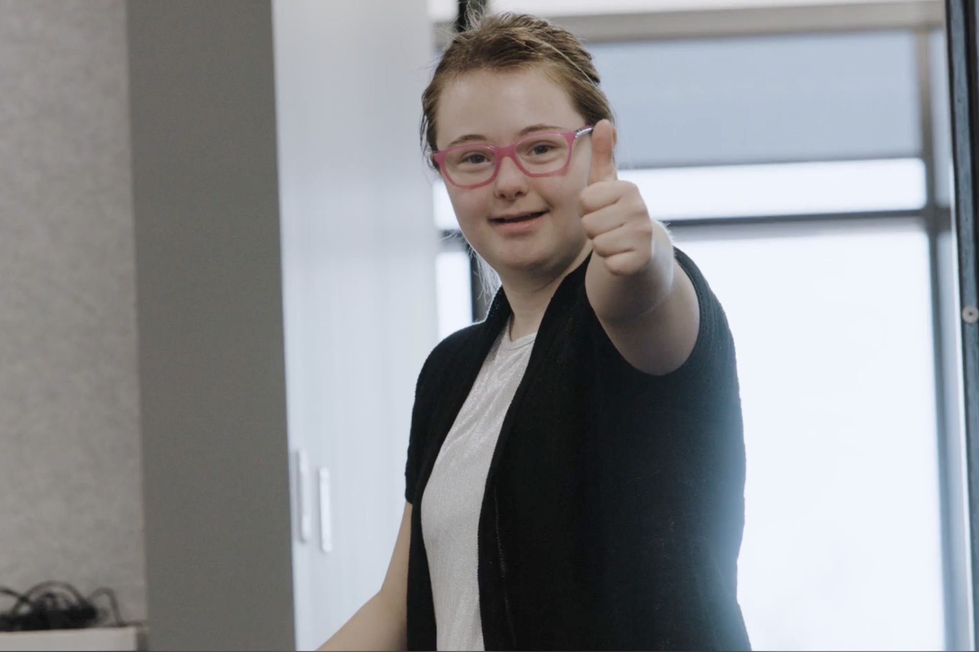 Person standing indoors giving a thumbs-up gesture, wearing a white shirt and dark cardigan, with a modern office background featuring gray walls and a window.