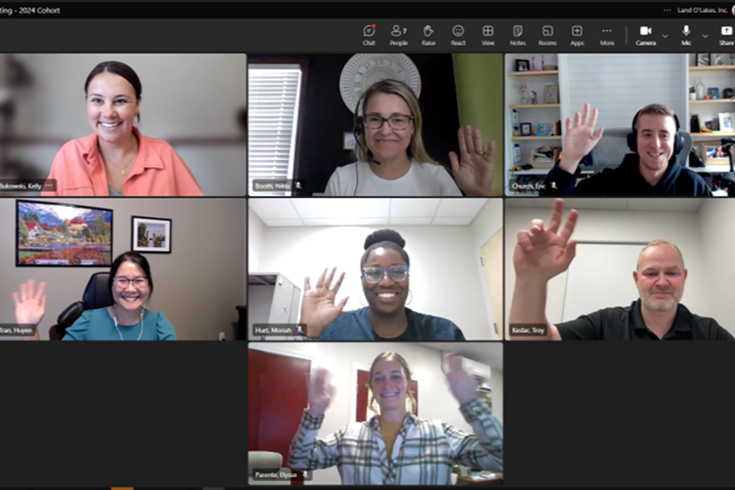 A group of employees seated on indoor bleachers in two rows, wearing business‑casual attire, with arena seating and a Land O’Lakes Inc. banner behind them.