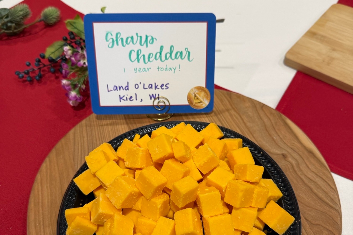 Plate of cubed sharp cheddar cheese on a wooden board with a sign reading “Sharp Cheddar — 1 year today! Land O’Lakes, Kiel, WI.”