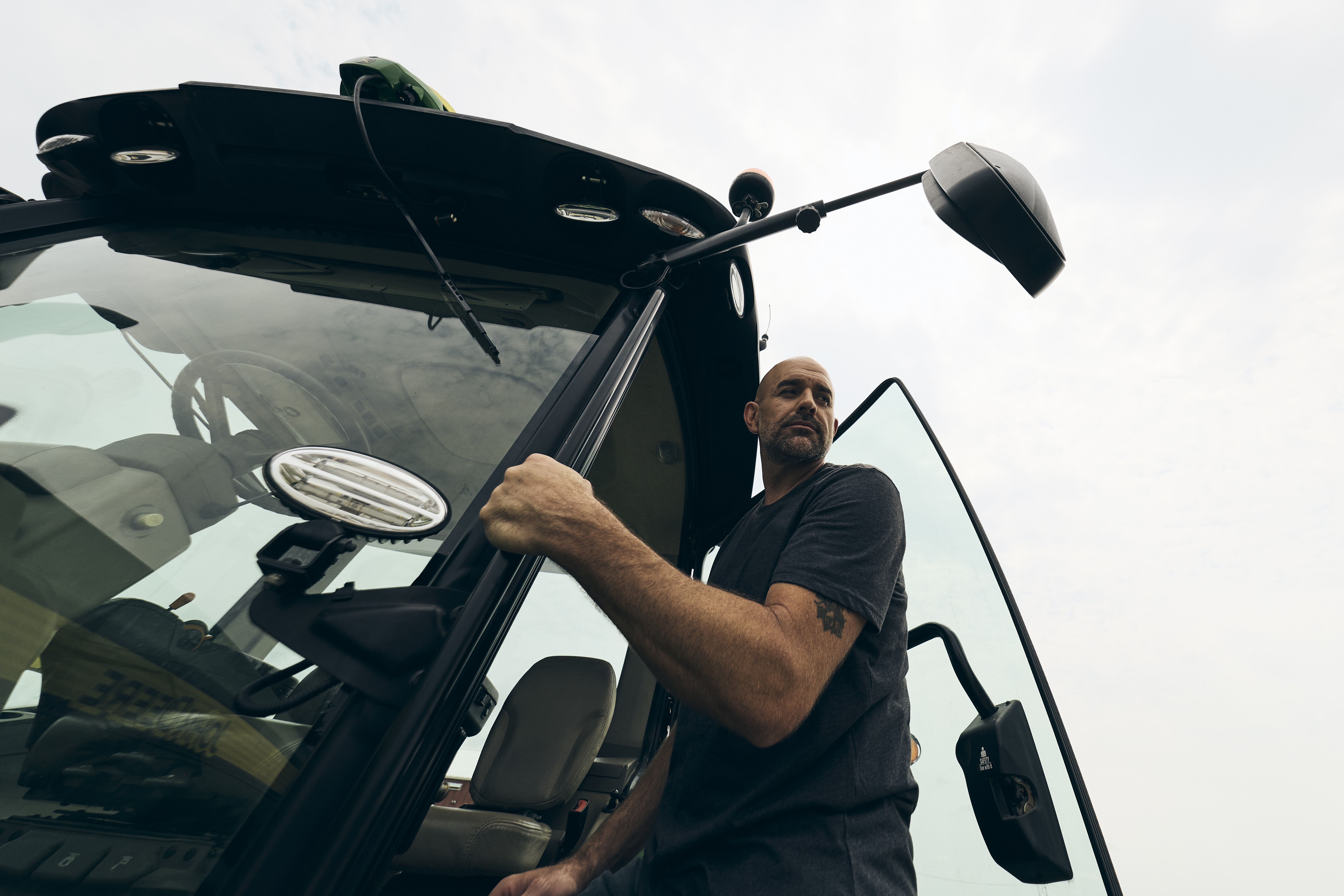 Low-angle view of a person climbing out of a tractor cab on a farm under a cloudy sky.