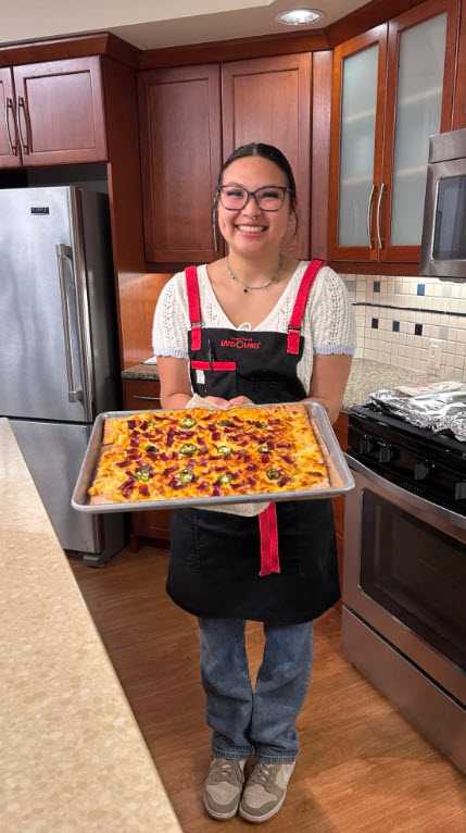 A Culinary Center intern wearing a Land O’Lakes apron holds a freshly baked sheet-pan pizza in a test kitchen with stainless steel appliances and wood cabinets.