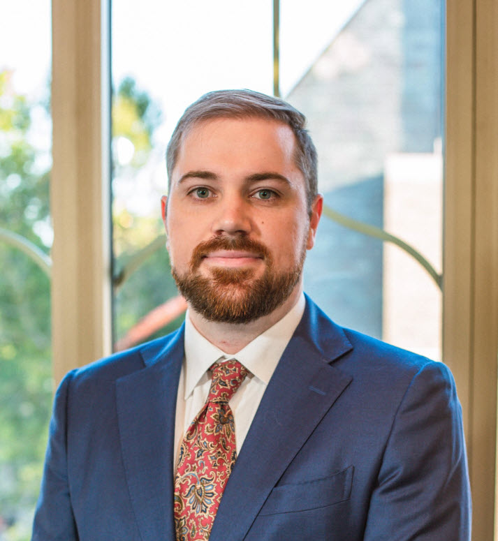 Professional headshot of a person in a blue suit and patterned tie standing in front of large windows.